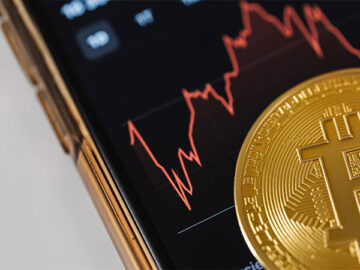 close up view of a gold bitcoin on top of a smartphone with line graph on its screen